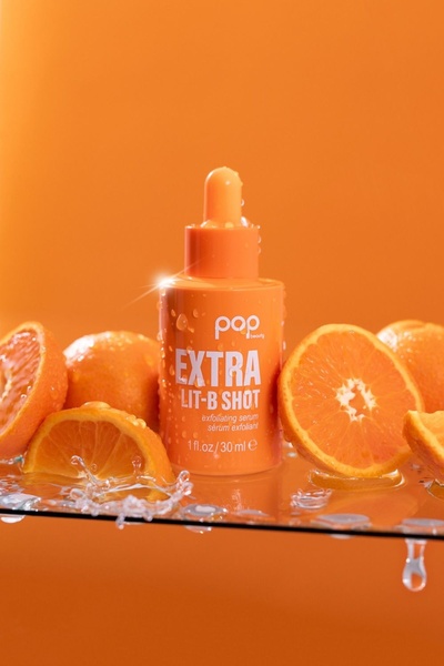 A high-energy, citrus-themed product shot showcasing an exfoliating serum bottle surrounded by fresh orange slices and water droplets.